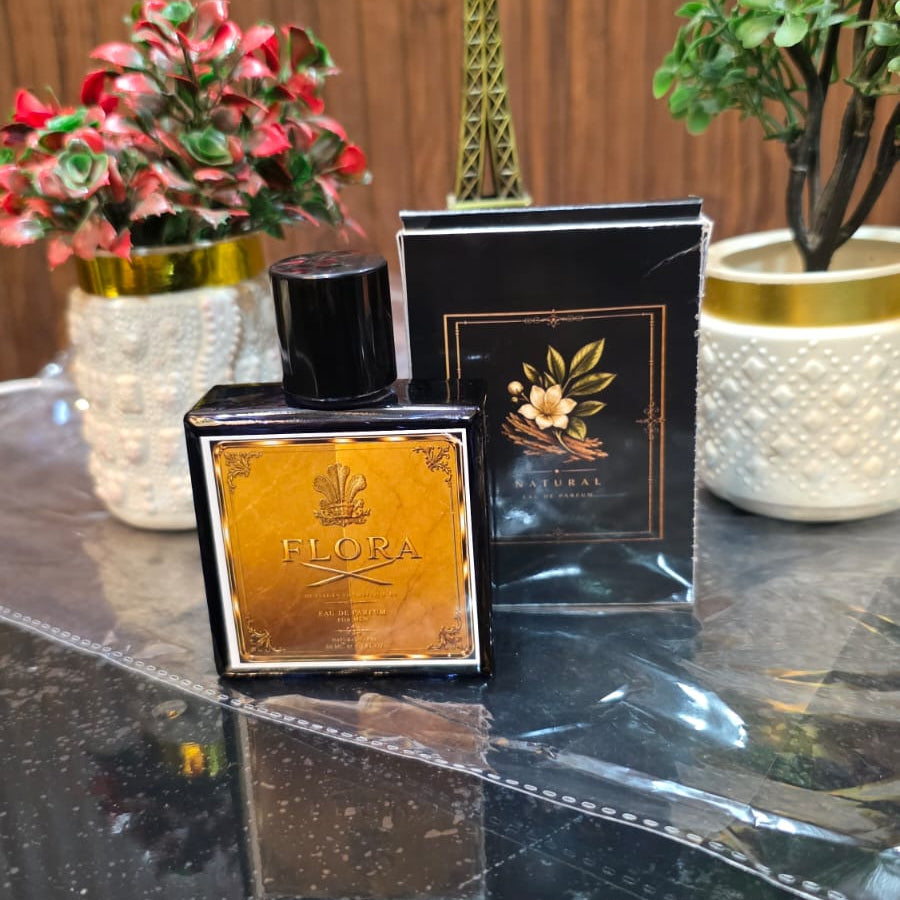 Two black perfume bottles with gold accents on a reflective surface, surrounded by decorative plants.